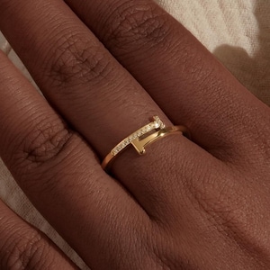 Peut inclure: Une bague dorée au design unique, avec une petite barre angulaire et un anneau orné de minuscules pierres scintillantes. La bague est portée sur une main, sur un fond neutre.