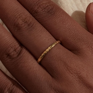 Könnte beinhalten: Ein goldener Ring mit strukturierter Oberfläche wird an einem Finger getragen. Das Design des Rings weist eine Reihe kleiner, sich wiederholender Muster auf. Der Ring ist vor einem neutralen Hintergrund platziert, was seine schlichte Eleganz unterstreicht. Der Ring ist ein zarter Schmuck.