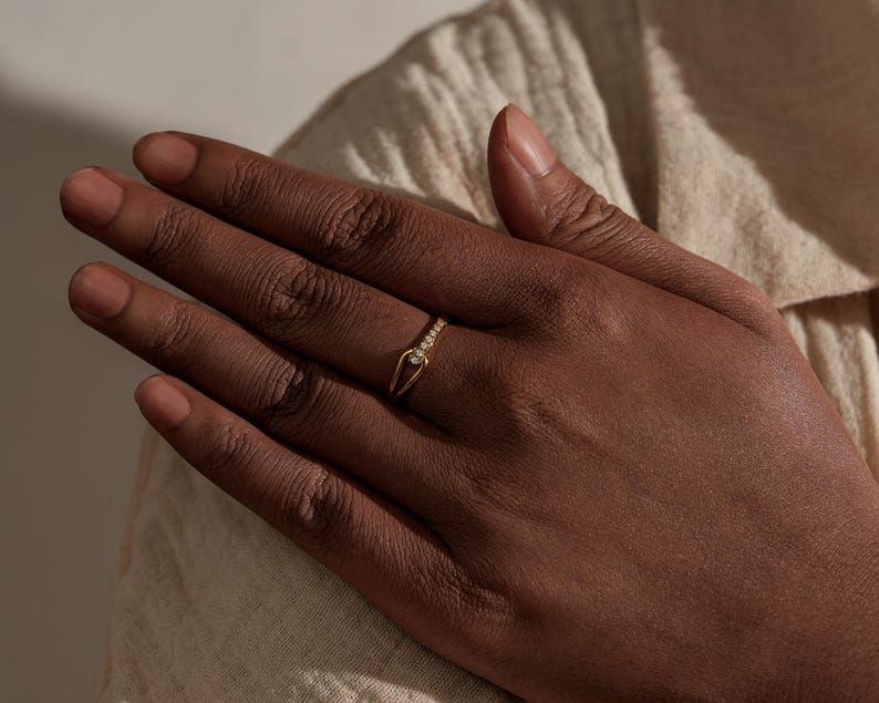 May include: A gold ring with a unique design, featuring a row of small, clear stones along the top band. The ring has a distinctive open shape with a pointed element. The ring is worn on a hand with dark skin, against a neutral background.