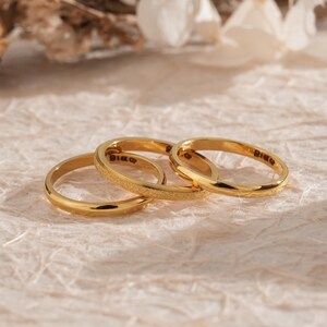 May include: Three gold rings are arranged on a textured, light-coloured surface. The rings have a simple design, with one ring featuring a textured surface. The rings are likely for engagement or wedding purposes. The rings are made of gold.