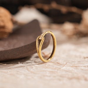 May include: A gold ring with a unique design, featuring a curved band and a small section adorned with tiny, sparkling stones. The ring is set against a neutral background, highlighting its elegant form and intricate details. Jewelry.