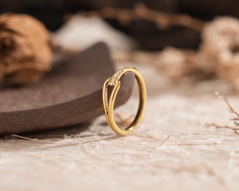 May include: A gold ring with a unique design, featuring a curved band and a small section adorned with tiny, sparkling stones. The ring is set against a neutral background, highlighting its elegant form and intricate details. Jewelry.