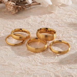 May include: A collection of gold-colored rings of varying widths and styles. The rings are arranged on a textured, off-white surface, with some overlapping. The lighting creates highlights and shadows, emphasizing the metallic sheen of the jewelry.