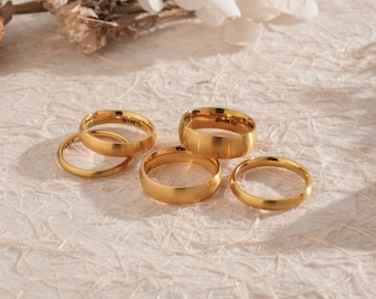 Könnte beinhalten: Eine Sammlung goldfarbener Ringe in verschiedenen Breiten und Stilen. Die Ringe sind auf einer strukturierten, cremefarbenen Oberfläche angeordnet, wobei sich einige überlappen. Die Beleuchtung erzeugt Glanzlichter und Schatten, die den metallischen Glanz des Schmucks hervorheben.