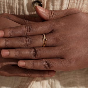 May include: A gold ring with a V-shaped design and small, sparkling stones. The ring is on a finger, with the hand resting on a cream-colored fabric. The ring's delicate design is highlighted against the skin tone.