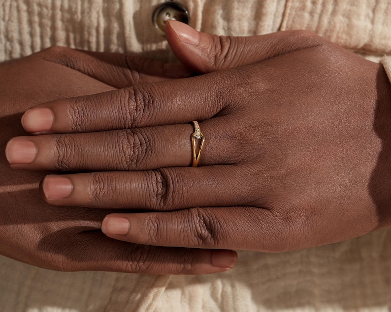 May include: A gold ring with a V-shaped design and small, sparkling stones. The ring is on a finger, with the hand resting on a cream-colored fabric. The ring's delicate design is highlighted against the skin tone.