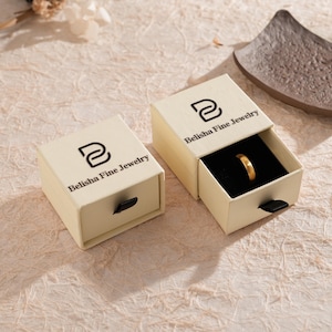 May include: Two beige jewelry boxes with the text "Belisha Fine Jewelry" printed on the top. One box is open, revealing a gold ring on a black interior. The boxes are on a textured, light-colored surface.