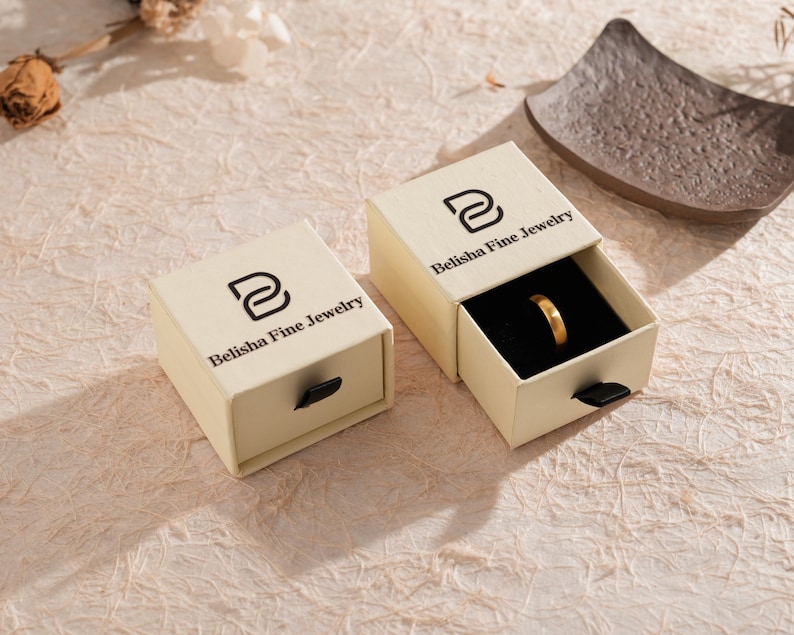 May include: Two beige jewelry boxes with the text "Belisha Fine Jewelry" printed on the top. One box is open, revealing a gold ring on a black interior. The boxes are on a textured, light-colored surface.