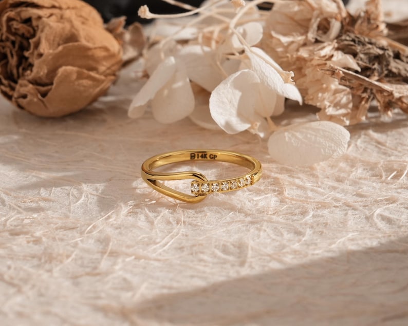 May include: A gold ring with a unique design, featuring a row of small, clear stones set into the band. The ring has a distinctive hook-like shape. The ring is set against a neutral background with dried flowers.