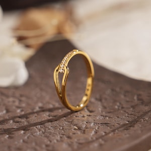 May include: A gold-colored ring with a unique design. The ring features a curved band with a small section adorned with small, clear stones. The ring is displayed on a brown surface, with soft lighting and a blurred background.