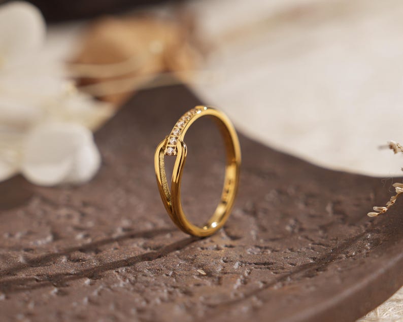 May include: A gold-colored ring with a unique design. The ring features a curved band with a small section adorned with small, clear stones. The ring is displayed on a brown surface, with soft lighting and a blurred background.