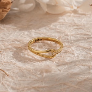 May include: A gold ring with a unique design. The ring features a curved band that intersects with a smaller band adorned with small, sparkling stones. The ring is set against a textured, off-white surface, with soft shadows and floral accents.