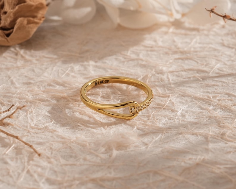May include: A gold ring with a unique design. The ring features a curved band that intersects with a smaller band adorned with small, sparkling stones. The ring is set against a textured, off-white surface, with soft shadows and floral accents.