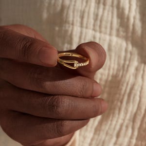 May include: A gold ring with a unique design, featuring a small row of clear stones and a curved band. The ring is held in a hand, showcasing its delicate details and elegant style. The ring appears to be made of gold.