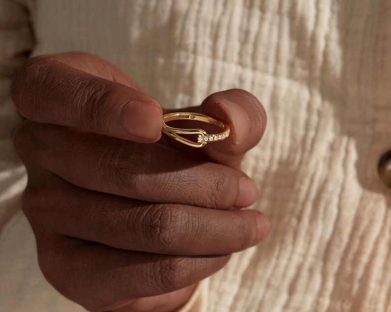 May include: A gold ring with a unique design, featuring a small row of clear stones and a curved band. The ring is held in a hand, showcasing its delicate details and elegant style. The ring appears to be made of gold.