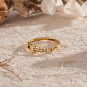 May include: A gold ring with a unique design. The ring features a series of small, clear stones set along one side. The band is a bright gold color, and the ring has a distinctive shape. The ring is set against a neutral background.