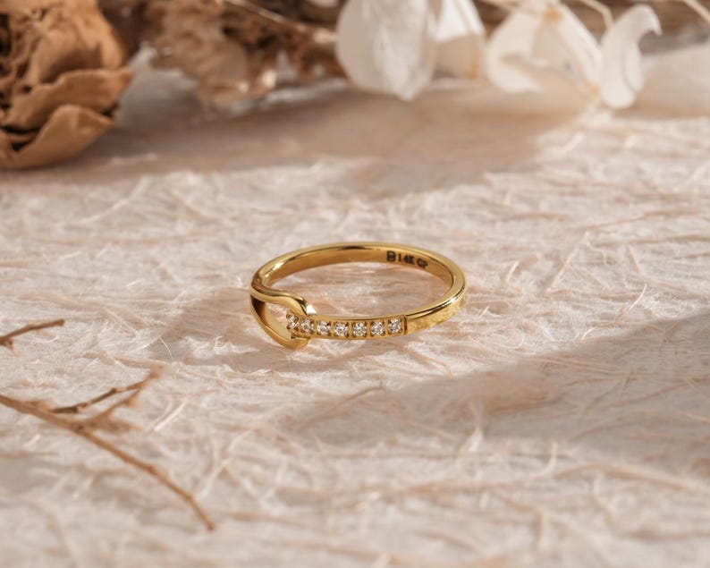 May include: A gold ring with a unique design. The ring features a series of small, clear stones set along one side. The band is a bright gold color, and the ring has a distinctive shape. The ring is set against a neutral background.