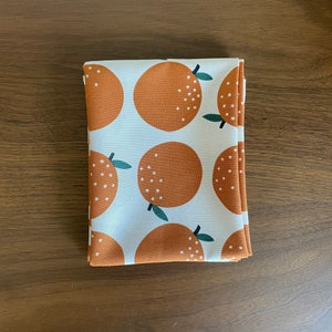 Könnte beinhalten: Weißer Stoff mit einem sich wiederholenden Muster aus orangefarbenen Zitrusfrüchten mit grünen Blättern. Die Orangen haben weiße Punkte.