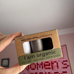 Puede incluir: Una caja de cartón rectangular que contiene tres carretes de hilo Scanfil Organics en blanco, gris y negro. El envase está etiquetado como 3 spools, i am organic, 50/2 WT y 500 YDS | 457 M EACH. La caja tiene una ventana que muestra el hilo.