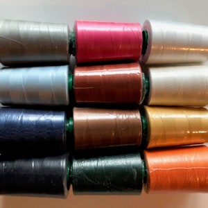 May include: A collection of 10 spools of thread in various colors, including gray, pink, white, light blue, brown, gold, orange, dark blue, green, and dark green. The spools are wrapped in clear plastic and have green caps.