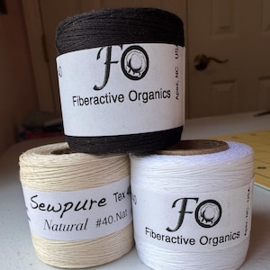 May include: Three spools of thread in black, natural, and white. The black spool is labelled "Fiberactive Organics." The natural spool is labelled "Sewpure Natural #40.Nat." The white spool is labelled "Fiberactive Organics."