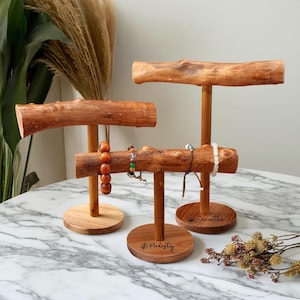 May include: Three wooden bracelet display stands with a natural wood finish. Each stand has a round base and a horizontal branch-like bar. Bracelets of various colours are displayed on the stands. The bases are engraved with names.