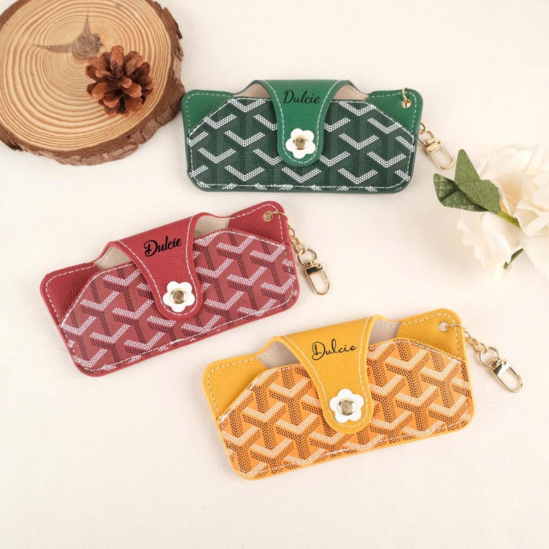 May include: Three eyeglass cases in green, red, and yellow, each with a geometric pattern and a white flower clasp. The word "Dulcie" is printed on the flap of each case, which also has a gold-colored keychain attachment.