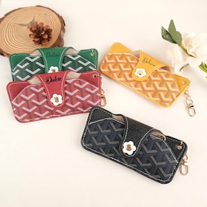 May include: Four eyeglass cases in green, red, yellow, and navy blue. Each case features a geometric pattern, a white flower-shaped clasp, and the word "Dulcie" in script. The cases have a gold-colored keychain attachment.