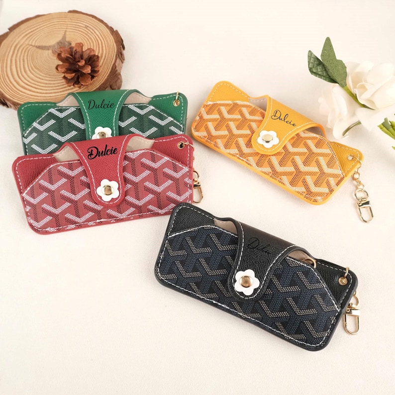 May include: Four eyeglass cases in green, red, yellow, and navy blue. Each case features a geometric pattern, a white flower-shaped clasp, and the word "Dulcie" in script. The cases have a gold-colored keychain attachment.