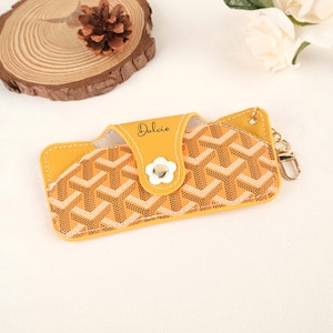 May include: Yellow eyeglass case with a geometric pattern in beige and white. The case has a snap closure with a white flower-shaped button. The word "Dulcie" is written in cursive on the flap. A gold-colored keychain is attached to the side.