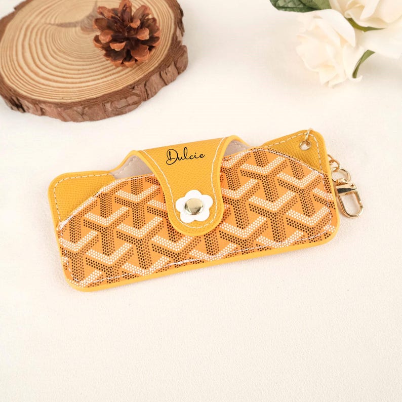 May include: Yellow eyeglass case with a geometric pattern in beige and white. The case has a snap closure with a white flower-shaped button. The word "Dulcie" is written in cursive on the flap. A gold-colored keychain is attached to the side.