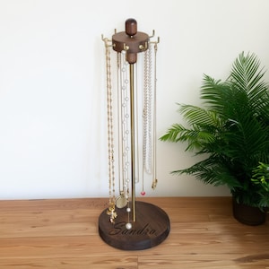 May include: A wooden jewelry stand with a dark brown base and top, connected by a brass-colored pole. The stand holds various necklaces of different styles and lengths. The base is engraved with the name "Sandra".