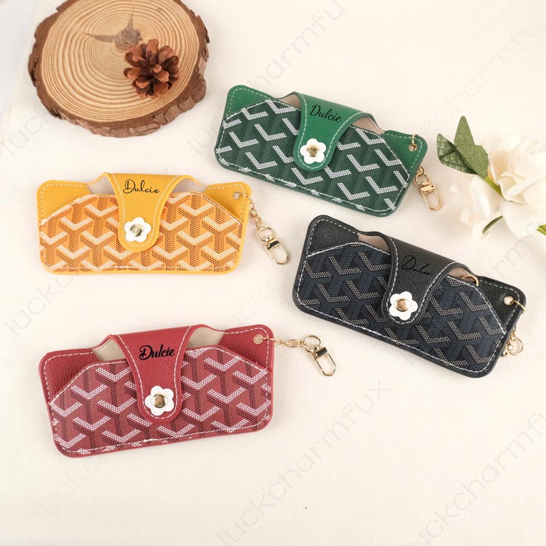 May include: Four leather eyeglass cases in yellow, green, black, and red. Each case features a geometric pattern, a white flower-shaped clasp, and the word "Dulcie". Gold-colored keychains are attached.
