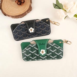 May include: Two cardholders with a geometric pattern. One is navy blue and the other is green, both with white accents and a flower-shaped clasp. The word "Dulcie" is printed on the flap of each cardholder.
