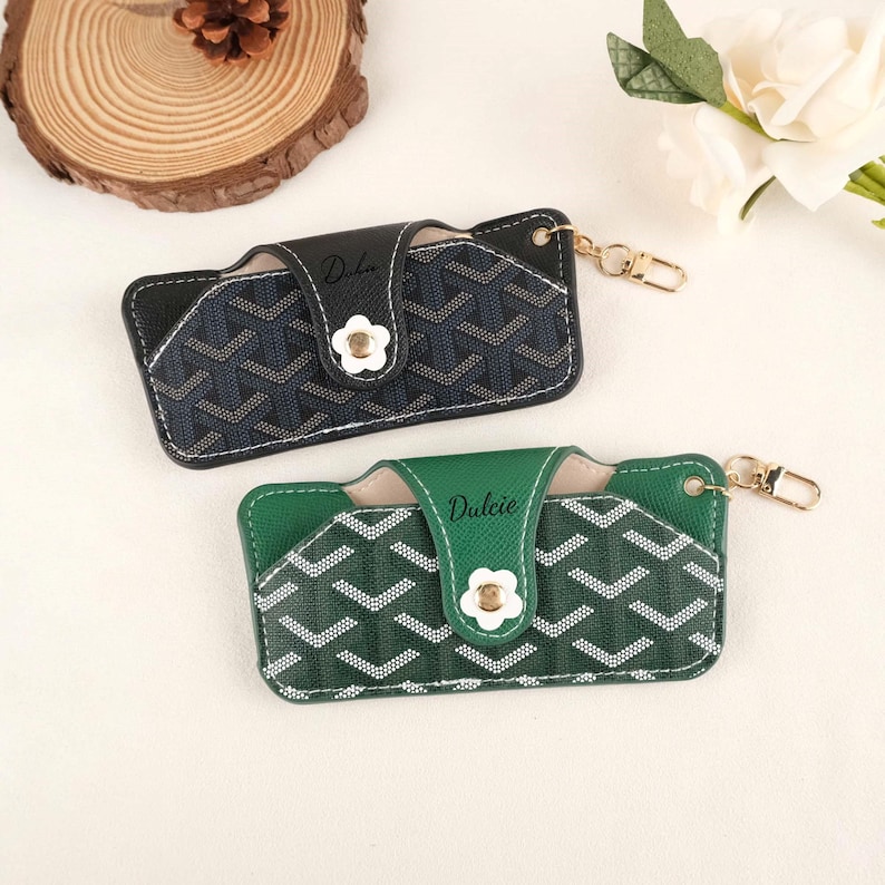 May include: Two cardholders with a geometric pattern. One is navy blue and the other is green, both with white accents and a flower-shaped clasp. The word "Dulcie" is printed on the flap of each cardholder.