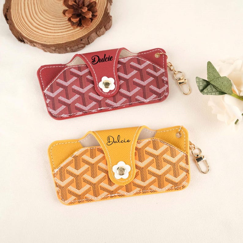 May include: Two leather eyeglass cases, one red and one yellow, with a geometric pattern. Each case has a flap with the name "Dulcie" and a flower-shaped button. They include a gold-colored keychain.
