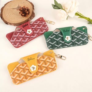 May include: Three leather eyeglass cases in red, green, and yellow. Each case features a geometric pattern, a small flower clasp, and the name "Dulcie" printed on the flap. Gold-colored keychains are attached.