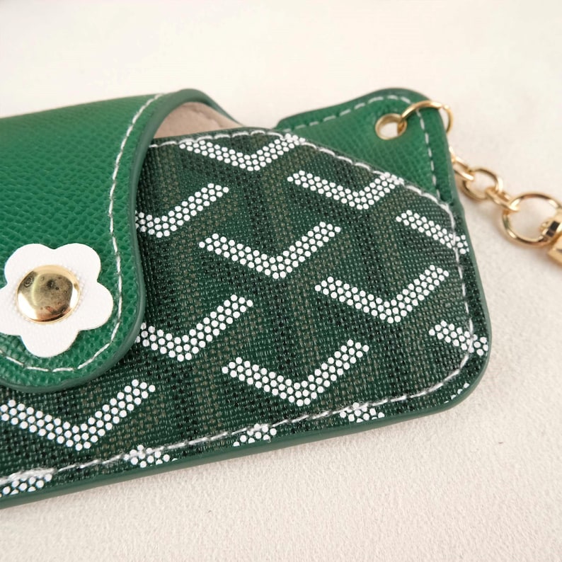 May include: A green key holder with a white flower-shaped clasp and gold-tone hardware. The key holder features a patterned fabric with white chevron designs. The stitching is white, and a gold chain is attached.