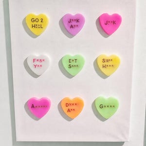 May include: Nine colorful heart-shaped candies with various phrases, including "GO 2 HEL" and "J**K", are arranged on a white background. The candies are in shades of yellow, pink, white, green, and orange.