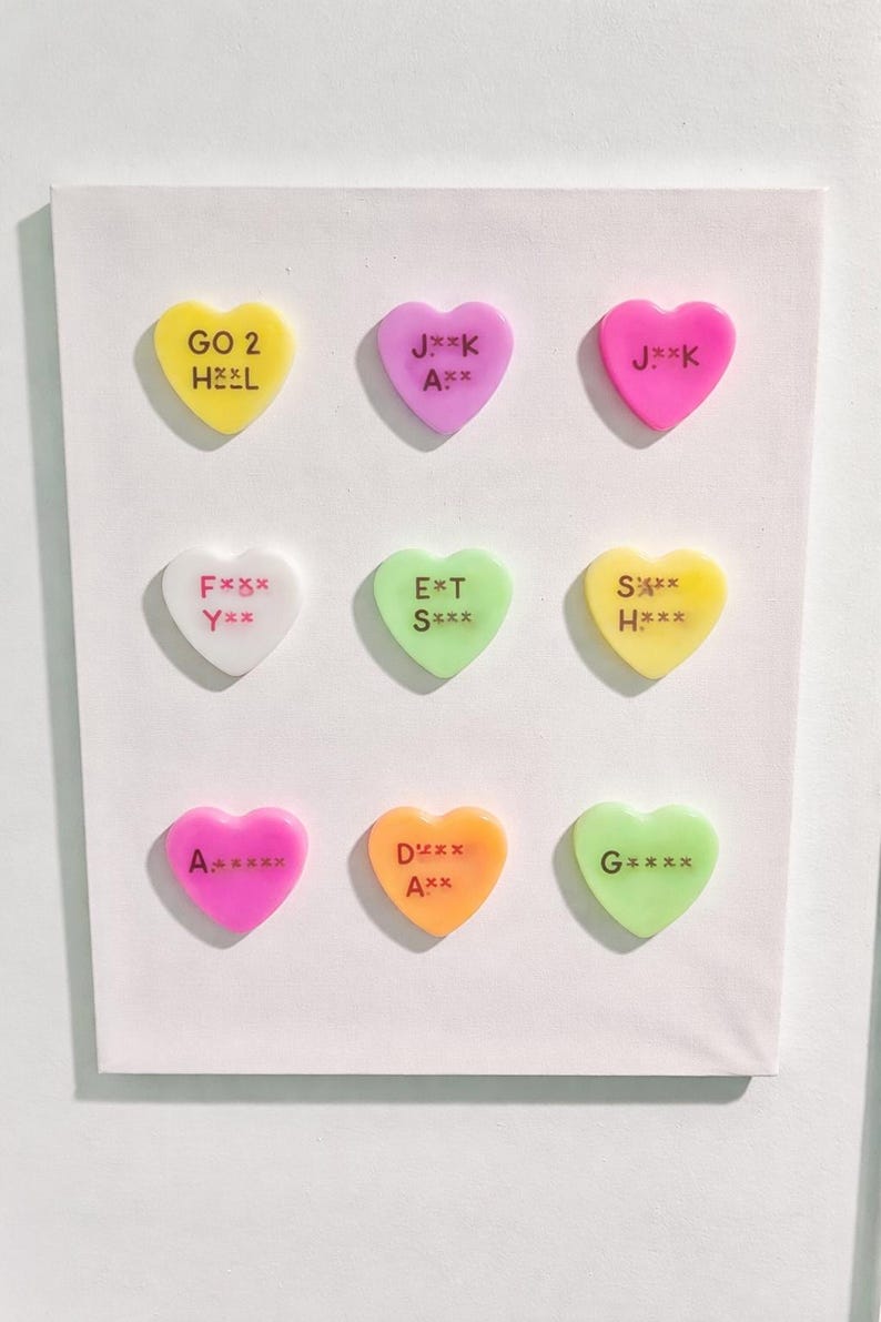 May include: Nine colorful heart-shaped candies with various phrases, including "GO 2 HEL" and "J**K", are arranged on a white background. The candies are in shades of yellow, pink, white, green, and orange.