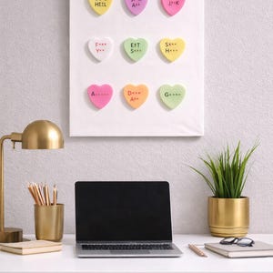 May include: A white canvas with nine colorful heart-shaped candies, each with a unique message. The desk setup includes a laptop, a gold lamp, pencils, and a potted plant. The candies have messages like "GO 2 HEZL" and "F*** Y**".