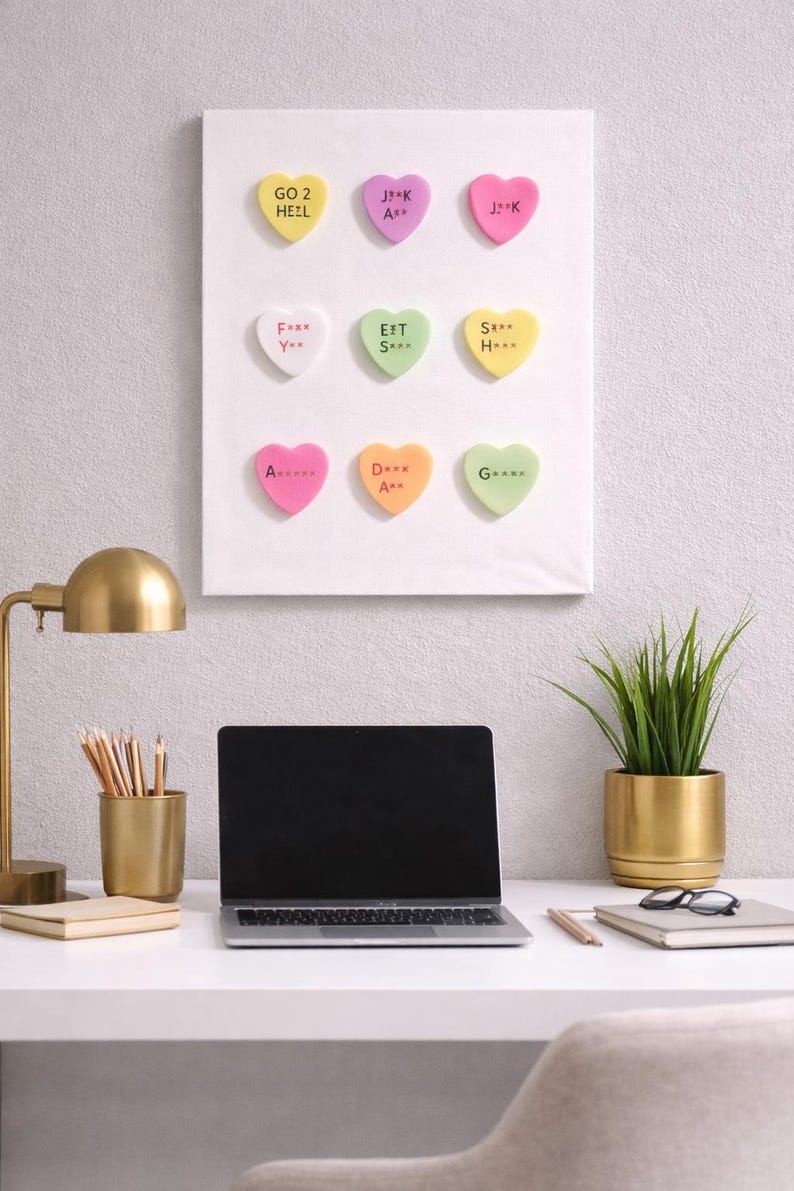 May include: A white canvas with nine colorful heart-shaped candies, each with a unique message. The desk setup includes a laptop, a gold lamp, pencils, and a potted plant. The candies have messages like "GO 2 HEZL" and "F*** Y**".
