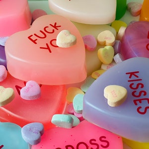May include: An assortment of heart-shaped candies and soaps in various colors, including pink, purple, and yellow. Some of the candies have messages like "Kisses" and "Fuck You" printed on them. The image has a close-up perspective.