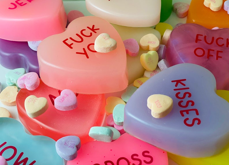 May include: An assortment of heart-shaped candies and soaps in various colors, including pink, purple, and yellow. Some of the candies have messages like "Kisses" and "Fuck You" printed on them. The image has a close-up perspective.
