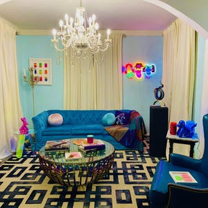 May include: A living room with a blue sofa, a patterned rug, and a glass-topped coffee table. A chandelier hangs from the ceiling, and a neon sign is on the wall. A blue armchair and a unicorn head are also visible.