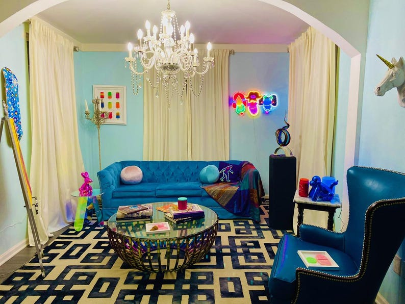 May include: A living room with a blue sofa, a patterned rug, and a glass-topped coffee table. A chandelier hangs from the ceiling, and a neon sign is on the wall. A blue armchair and a unicorn head are also visible.