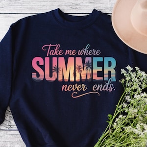 Op de afbeelding: Marineblauw sweatshirt met de tekst "Take me where SUMMER never ends." Het woord "SUMMER" is in een verloop van roze, oranje en blauw, met silhouetten van palmbomen. Een beige hoed en bloemen staan op de achtergrond.