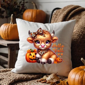 Puede incluir: Una almohada blanca con una vaca de dibujos animados linda que lleva una diadema y gafas de leopardo marr&oacute;n y negro. La vaca sostiene una peque&ntilde;a calabaza naranja y la almohada dice "Hello Fall" en texto negro.
