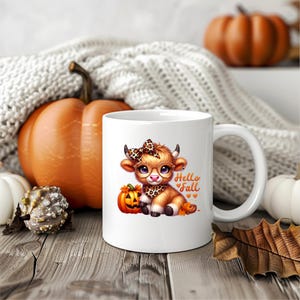 Puede incluir: Taza de cer&aacute;mica blanca con una ilustraci&oacute;n de dibujos animados de una vaca marr&oacute;n y blanca con gafas y un lazo. La vaca sostiene una peque&ntilde;a calabaza naranja con una cara de calabaza. El texto "Hello Fall" est&aacute; impreso en la taza en negro.