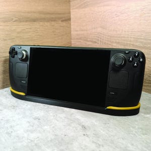 May include: A black handheld gaming device with yellow accents. The device has a large screen, two joysticks, and a variety of buttons. The device is sitting on a grey surface.